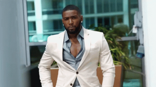 A Man In A White Suit And Blue Shirt Is Standing In Front Of A Building GIF