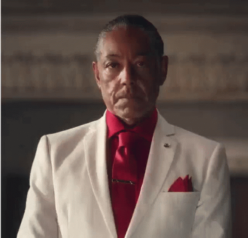 A Man In A White Suit And Red Tie Looks At The Camera GIF