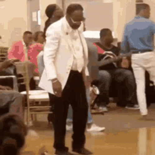 A Man In A White Suit Is Dancing In Front Of A Crowd Of People In A Room . GIF