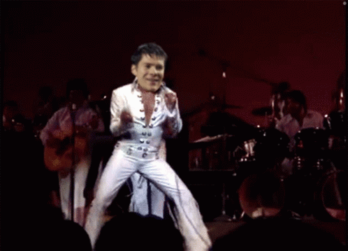 A Man In A White Suit Is Dancing On A Stage In Front Of A Band GIF