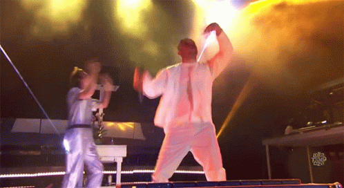 A Man In A White Suit Is Dancing On A Stage While A Woman Stands Behind Him With A Sign That Says Just Dance GIF