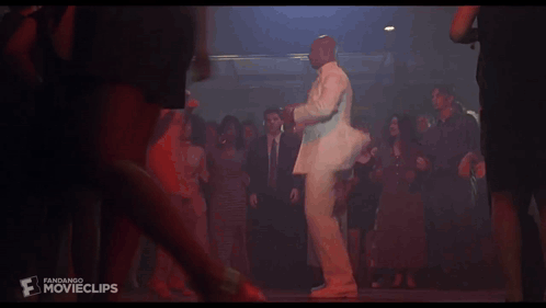 A Man In A White Suit Is Dancing With A Woman In An Orange Dress At A Party GIF