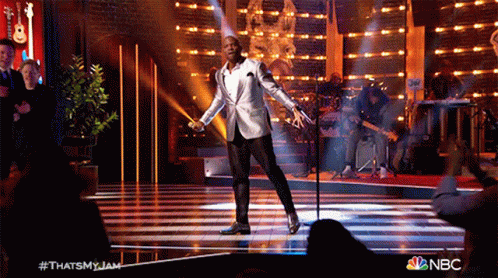 A Man In A White Suit Is Singing Into A Microphone On A Stage With A Nbc Logo In The Corner GIF