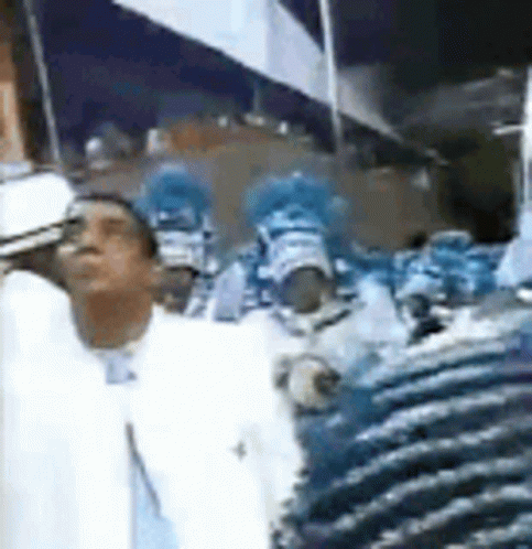 A Man In A White Suit Is Standing In Front Of A Crowd Of People In Blue Costumes . GIF