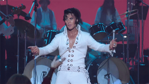 A Man In A White Suit Is Standing In Front Of A Drum Set On A Stage GIF