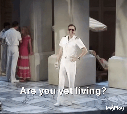 A Man In A White Suit Is Standing In Front Of A Group Of People And Says Are You Yet Living GIF