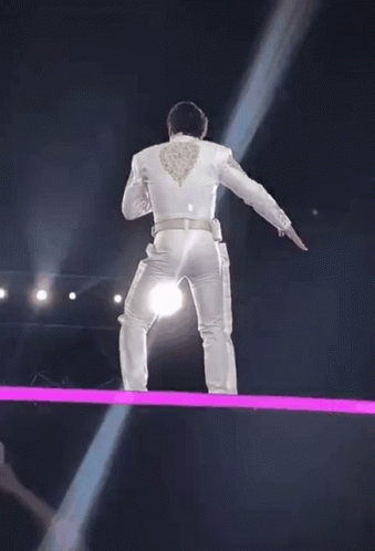 a man in a white suit is standing on a stage with a light coming from his back .