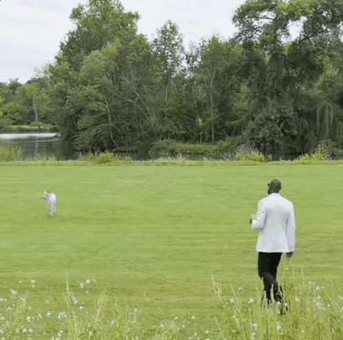A Man In A White Suit Is Walking A Dog In A Field GIF