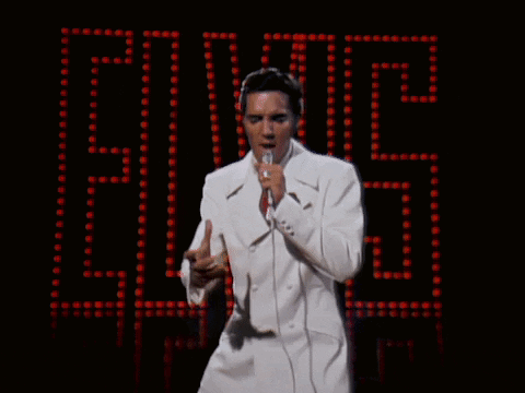 A Man In A White Suit Sings Into A Microphone In Front Of A Elvis Sign GIF