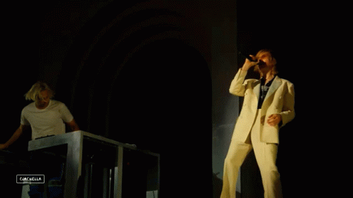A Man In A White Suit Sings Into A Microphone While Another Man Plays A Keyboard In The Background GIF