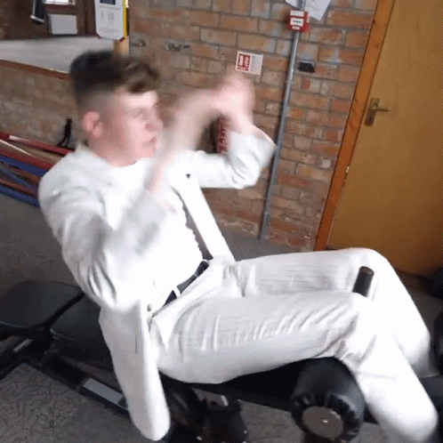 A Man In A White Suit Sits On A Bench With A Fire Extinguisher On The Wall Behind Him GIF