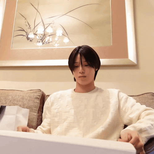 A Man In A White Sweater Is Sitting On A Couch In Front Of A Picture GIF