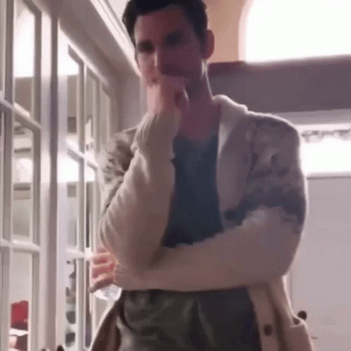 A Man In A White Sweater Is Standing In A Room With His Arms Crossed And His Hand On His Chin . GIF