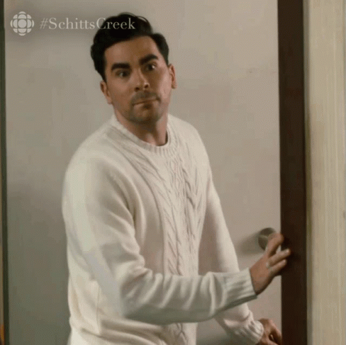 A Man In A White Sweater Is Standing In Front Of A Door . GIF