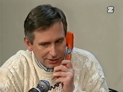 A Man In A White Sweater Is Talking On A Red Telephone GIF