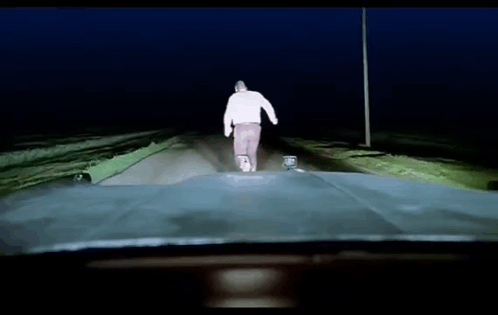 A Man In A White Sweater Is Walking Down A Road At Night GIF
