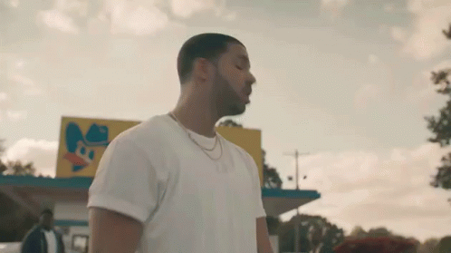 A Man In A White T-Shirt And Necklace Is Standing In Front Of A Fast Food Restaurant . GIF