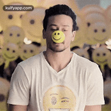 A Man In A White T-Shirt Is Blowing A Yellow Ball With A Smiley Face On His Nose . GIF