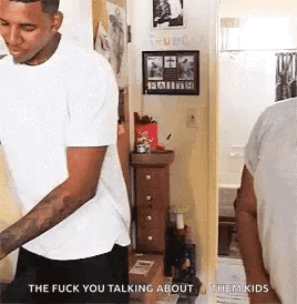 A Man In A White T-Shirt Is Standing In A Hallway Talking To Another Man . GIF