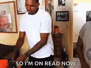 A Man In A White T-Shirt Is Standing In A Living Room Holding A Box . GIF