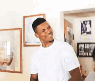 A Man In A White T-Shirt Is Standing In A Living Room With A Smile On His Face . GIF