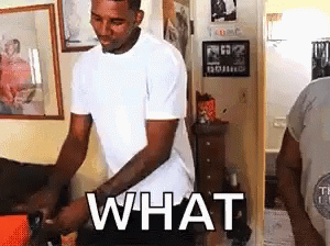 A Man In A White T-Shirt Is Standing In A Living Room With The Word What Written On His Chest . GIF