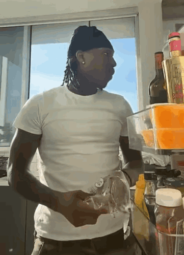 A Man In A White T-Shirt Is Standing In Front Of A Fridge Filled With Bottles And Condiments GIF