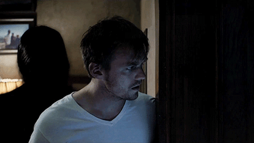 A Man In A White T-Shirt Stands In A Dark Room GIF