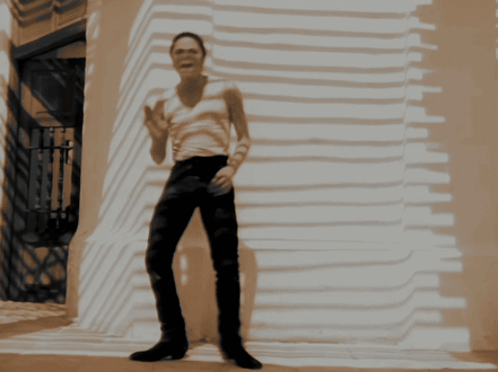 a man in a white tank top and black pants is standing in front of a building