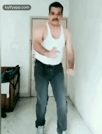 A Man In A White Tank Top And Blue Jeans Is Dancing In A Room . GIF