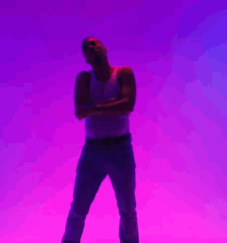 A Man In A White Tank Top And Blue Jeans Is Standing With His Arms Crossed In Front Of A Purple Background GIF