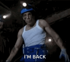 A Man In A White Tank Top And Blue Shorts Is Standing In A Dark Room And Says `` I 'M Back '' GIF