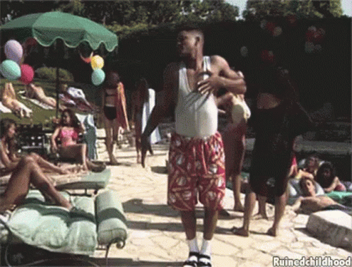 A Man In A White Tank Top And Red Shorts Is Standing In Front Of A Group Of People Near A Pool .. GIF