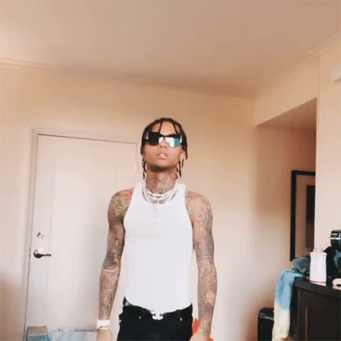 A Man In A White Tank Top And Sunglasses Stands In A Room GIF