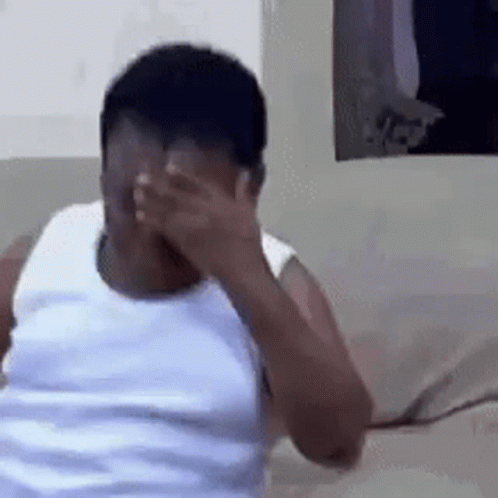 A Man In A White Tank Top Is Covering His Face With His Hand While Sitting On A Couch . GIF