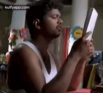 A Man In A White Tank Top Is Holding A Book In His Hands And Looking At It . GIF