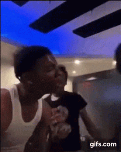 A Man In A White Tank Top Is Kissing Another Man On The Cheek In A Room . GIF