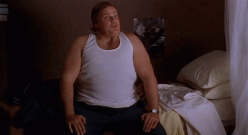 A Man In A White Tank Top Is Sitting On A Bed . GIF