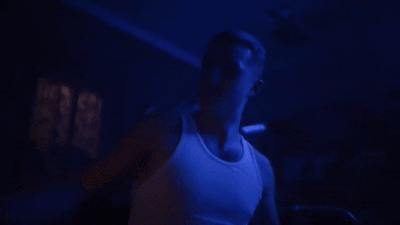 a man in a white tank top is standing in a dark room with blue lights behind him .