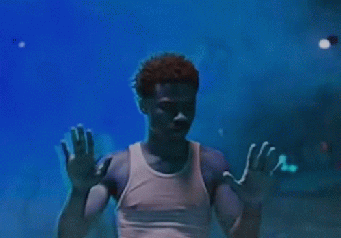 A Man In A White Tank Top Is Standing In A Dark Room With His Hands Up In The Air . GIF