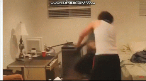 a man in a white tank top is standing in a room with a computer on the desk .