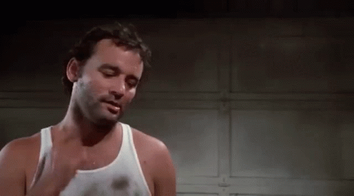 A Man In A White Tank Top Is Standing In Front Of A Garage Door And Smoking A Cigarette . GIF