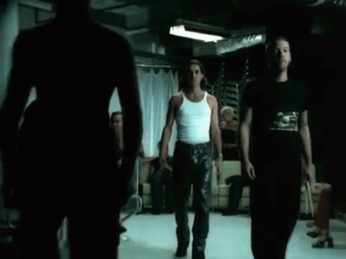 A Man In A White Tank Top Is Standing In Front Of A Group Of People In A Dark Room . GIF