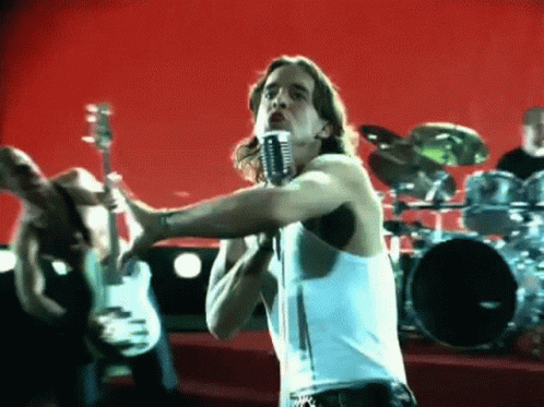 A Man In A White Tank Top Singing Into A Microphone In Front Of A Drum Set . GIF