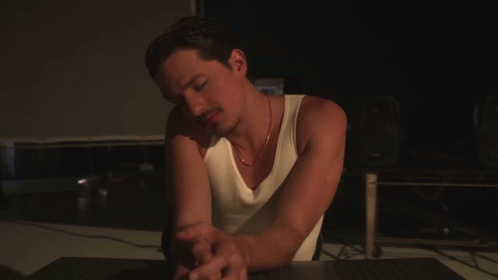 A Man In A White Tank Top Sits At A Desk GIF