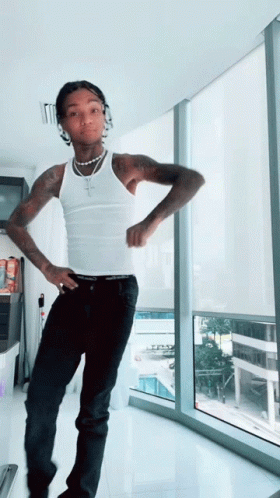 A Man In A White Tank Top Stands In Front Of A Large Window GIF