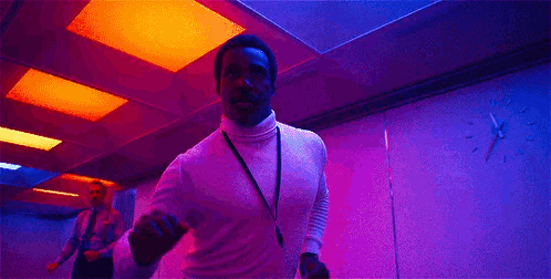 a man in a white turtleneck is dancing in a pink room with a lanyard around his neck .