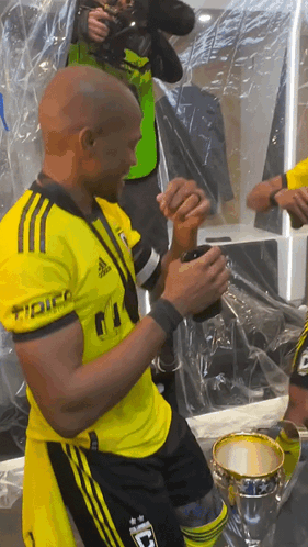 A Man In A Yellow Adidas Shirt Holds A Cup In His Hand GIF