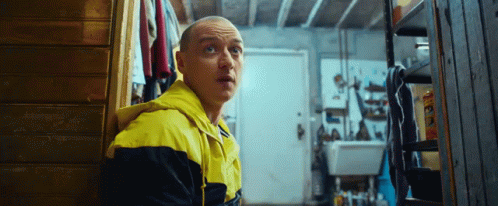 A Man In A Yellow And Black Jacket Is Standing In A Room Looking At Something . GIF