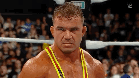 A Man In A Yellow And Red Tank Top Is Standing In A Wrestling Ring Looking At The Camera . GIF